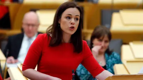 Getty Images Kate Forbes addresses MSPs at a wooden podium inside the Scottish parliament  debating chamber, wearing a bright red dress. The background features rows of wooden seats and other MSPs seated.