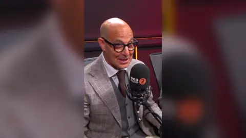 Stanley Tucci is wearing a grey suit with a brown tie and glasses. He is sitting in front of a BBC Radio 2 microphone.