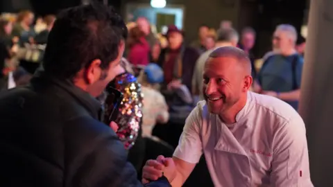 La Vida Liverpool & Jade Andreas Dave Critchley wears a chef coat with his name embroidered on the chest. He is shaking hands with another man and smiling. 