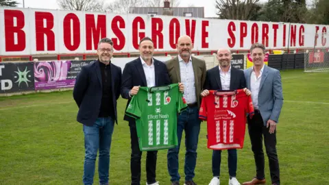 Five men wearing smart jackets, shirts and jeans hold up two football shirts on a football pitch with a white board behind them that reads "Bromsgrove Sporting" in red lettering.