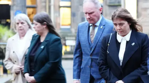 PA Media Ethan Scott Brown's mother Tracy Scott (right) and step-father Colin Scott with other two family members in the background. Tracy has shoulder-length brown hair and is wearing a navy jacket over a blouse with a white collar. Colin has short receding grey hair and is wearing a blue suit with a pale stripe. They are both looking to the ground.