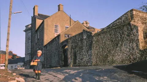 Armagh County Museum A digitalised photo from a film of an old stone building. A lady wearing a navy coat can be seen walking past. 
Looking north east from Callan street to Armagh Public Library. A woman in blue raincoat carrying a shopping basket walks towards the camera in the evening sunshine