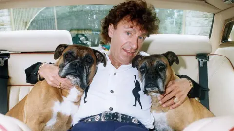 Getty Images Frank Pearson with brown curly hair wearing a white shirt and denims hugging two boxer dogs with each arm in the back of a car 