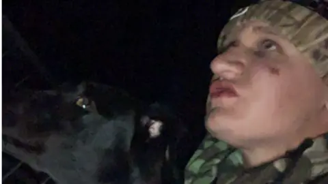 A man wearing a wooly hat and camouflaged top with a black dog. Both of them are staring off to the side of the camera