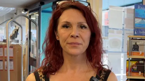 Nick Gaskell has wavy red hair. She has glasses resting on her head and is standing in front of the double doors to the library with a security scanner to the left and books to the right.