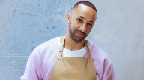 A man with short black hair and beard stands with his head cocked to one side. He is wearing a purple cardigan over a white jumper, with a brown apron over the top.