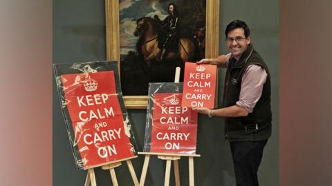 A dark-haired man standing next to two easels with red Keep Calm and Carry On posters on. He is also holding a smaller version of the poster.
