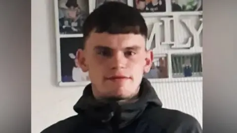 Handout A blurry head and shoulders shot of Kieran - he has dark hair and very short fringe and is wearing a black hoodie. There are lots of photos on the wall behind him and a white sign partially covered by his head with the letters - MILY visible - presumably for family. He is looking straight at the camera with a neutral expression on his face. 