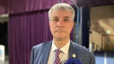 Sarah Sanderson / BBC A head and shoulders shot of councillor Philip Jackson who is looking glum. He has short grey hair and is wearing a grey checked blazer, a white shirt, a tie with small British flags on and a dark blue badge. 
