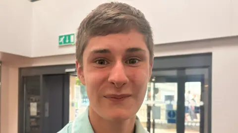 HTP Apprenticeship College Alfie Hobbs, a young man with short light brown hair, smiles for a posed picture. He is wearing a collared shirt with green stripes and is stood in front of the inside of an automatic doorway. 