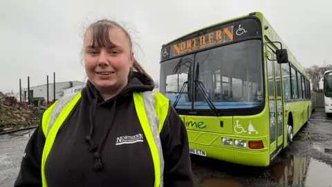 Rachel Smith, who is wearing a black hoodie with the Northern National Restoration Group's logo on and a high visibility jacket, stands in front of one of the remaining vehicles in the collection. It is a green bus with the words 'Lime' printed on the front. 