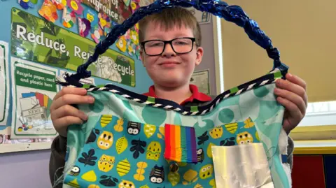 Cillian - a young child wearing black, square framed glasses holds up a large blue, leaf patterned bag with a blue, plaited handle.