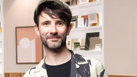 Designer John Cooper looks at the camera. He has a black beard and is wearing a tweed cap, monochrome shirt and black T-shirt