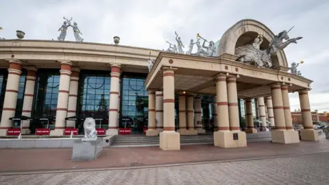The Trafford Centre The exterior of The Trafford Centre shopping centre, with large stone pillars and stone lions outside, and statues of angels ad a unicorn on the roof.
