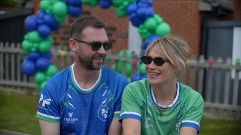 Connor Bennett/BBC A man and woman sit side by side, each wearing sunglasses and looking towards each other. He is on the left, has a brown hair and beard and wearing a blue football top. She has blonde hair tied back and is wearing a blue football top.