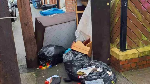 Hull City Council Three black bin bags dumped on a street, one with Lego spilling out of it. Two large brown wooden bedframes are dumped next to the bags, with one standing upright and one on its side