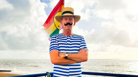 Fatboy Slim, Norman Cook, in a stripey sailor top stood with the sea at his back, a fake moustache on his face as he is staring at the camera with his arms folded and a hat on his head.