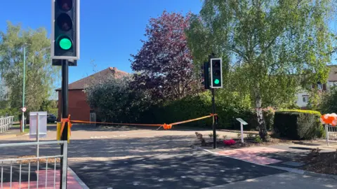 The crossing has an orange ribbon going from one side of the road to the other on the day of the official opening. Railings are to the side on the photo and bushes and trees are in the background.