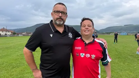Jak Powles who has brown hair and is wearing a red lions rugby shirt, with his arm around his father Carl, who has short dark hair, glasses and a beard and is wearing a black Port Talbot Panthers shirt. Behind them is a grass rugby pitch.