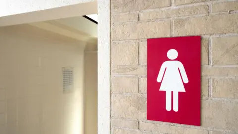 A generic photo of a ladies' toilet sign on a wall outside a toilet. The sign has a white lady symbol on a red background. The doorway has pale cream tiles.