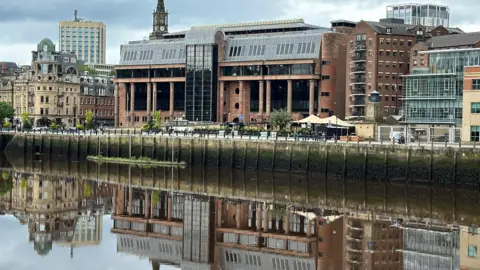 Newcastle Crown Court reflected on the River Tyne. It is a large imposing building of red bricks and columns with large dark windows.