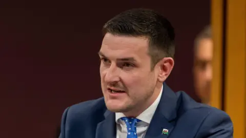 James Evans is wearing a blue suit, blue tie and white shirt, in front of a dark background. He is looking away from the camera.