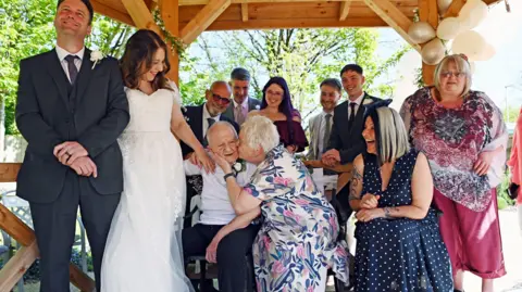 Richard Swingler Wedding celebrations including a bride and groom on the left and other family members wearing dresses and suits. The group are in some kind of wooden gazebo with trees seen in the background. 