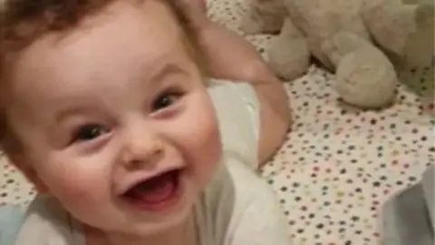 Police handout Close up of Preston's face as he laughs with his mouth open lying on his tummy in a cot with a spotty sheet. He is wearing a babygrow and the legs of a teddy can be seen to the top right
