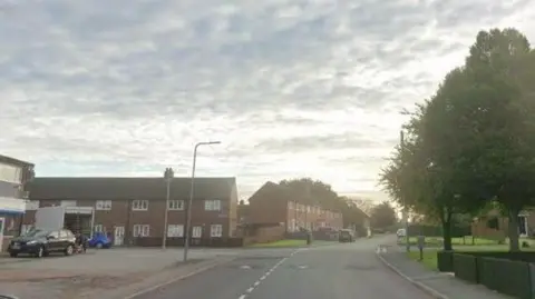 Google Sherlock Avenue in Haydock with shops on the left and trees on the right. Two blocks of two-storey terraced houses are in the distance at right angles to each other.