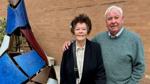 Woman with dark hair and black jacket stood next to man with grey hair and green jumper. They are stood next a cone-like art installation with blue and purple panels