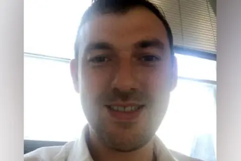 Hertfordshire County Council Gregory Assemakis: A man dark hair and stubble wearing an open-necked light shirt. 