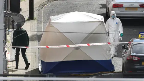 Margaret McLaughlin Man in forensics white suit beside a white tent, police tape and an officer pictured alongside scene