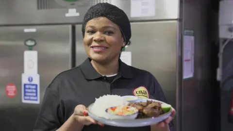 BBC Photograph of Justina John, owner of Onja restaurant. She wears a black polo top which reads "Onja, taste of Tanzania" on the yellow circle logo. Her hair is tied up into a black fishnet and she smiles at the camera. She holds a plate of food up to the camera.