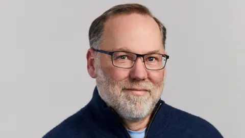 Fred Ramsdell, wearing a navy blue jumper and black-rimmed glasses, looks toward the camera in front of a neutral background.