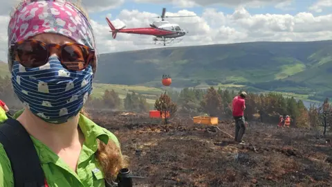 Peak District National Park Authority A selfie by a woman in protective clothing in front of a helicopter flying in a load of water to a burnt landscape