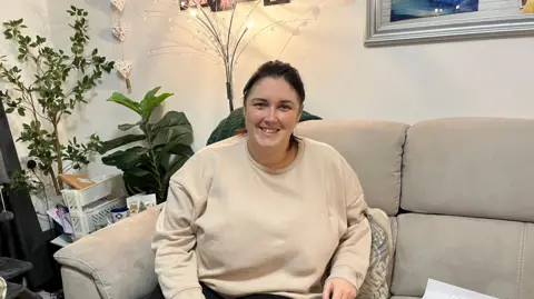 A woman in a cream jumper sitting on a grey sofa smiling. Behind her is a decorative lamp along with two house plants. There's also a coffee table to the left of her with a mug and various stationary bits tucked in.