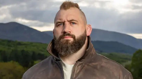 Joe Marler pictured wearing a jacket and looking stern with trees and mountains in the background
