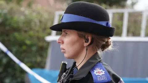 Essex Police A close-up side profile of a female police officer in a blue and black uniform.  She is standing outside at the scene of Richard Langley's alleged murder.