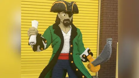 Captain Blackbrick. A model of a pirate made out of Lego-type bricks. The model depicts a pirate wearing blue trousers, a red belt, a white top, a green jacket with gold buttons and a black hat. He carries a sword in his left hand and a scroll in his right. 