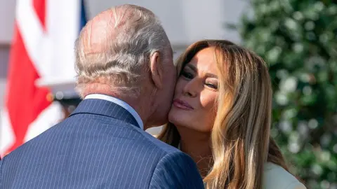 Getty Images King Charles in a pin stripe suit greets First Lady Melania with a kiss on the cheek during an arrival ceremony outside the White House in Washington. She has long hair. A union flag sits in the background.