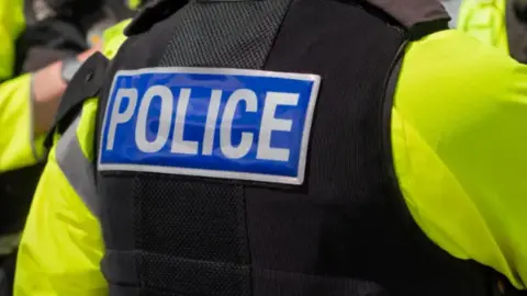 Getty Images A close up of the back of a police officer who is wearing a a high-viz jacket with a black stab vest over the top. The vest has a badge on it which reads "POLICE" in white on a blue background.