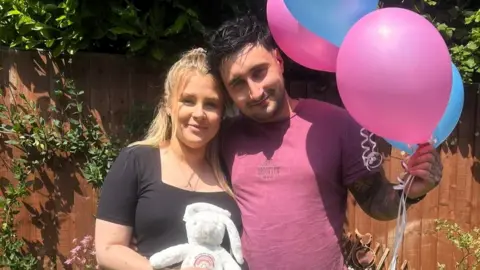 Oliver Dent Oliver Dent wearing a plum coloured t-shirt and holding pink and blue balloons. He is stood next to Caitlin who/s wearing a black t-shirt and holding a toy rabbit.