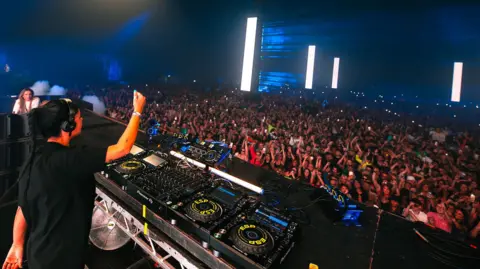 Josef Hall A woman with black hair tied up is wearing a black t-shirt and is DJing in front of a large crowd in a tent.