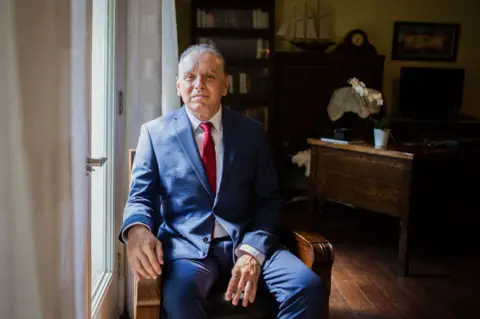 Dr Marrero, in a navy suit and red tie, sits in a wooden chair in his home office, in the light coming in from the door windows.