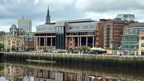 BBC Newcastle Crown Court reflected in the River Tyne running in front of it. It is an imposing building made from smooth red stone with massive black windows and tall columns along its frontage.