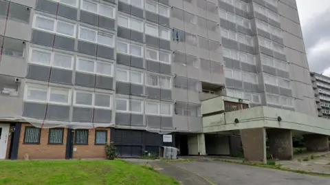 The tower block can be seen with a grey, concrete entrance on one side and a small grass area in front.