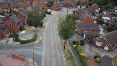 An aerial view of a suburban housing estate shows a wide residential road lined with detached and semi-detached brick homes. Several wooden telegraph poles stand alongside the road, with driveways, gardens and trees visible throughout the neighbourhood.