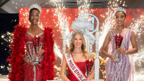 Miss England/Alan Strutt Three women all wearing sparkly dresses stand in front of pyrotechnics which are in full flow. Austin is on the left wearing a sparkly red dress, red feather boa and holding a trophy. In the middle sits a blonde woman on a throne who is wearing a Miss England sash and clutching a trophy. On the right, is a third woman wearing purple and clutching a trophy