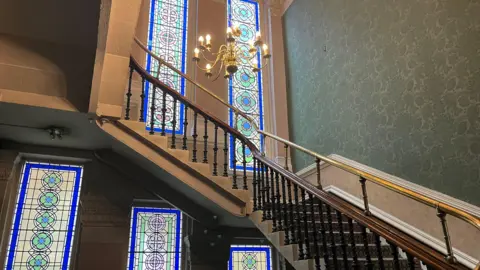 A staircase with a hanging light over it in front of stained glass windows