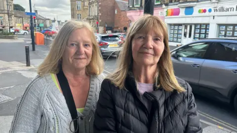 Jessica Bayley/BBC Two women stand together on a town street, the oney both have shoulder length blonde hair. The one on the left is wearing a grey cardigan, the one on the right is wearing a black waterproof coat.